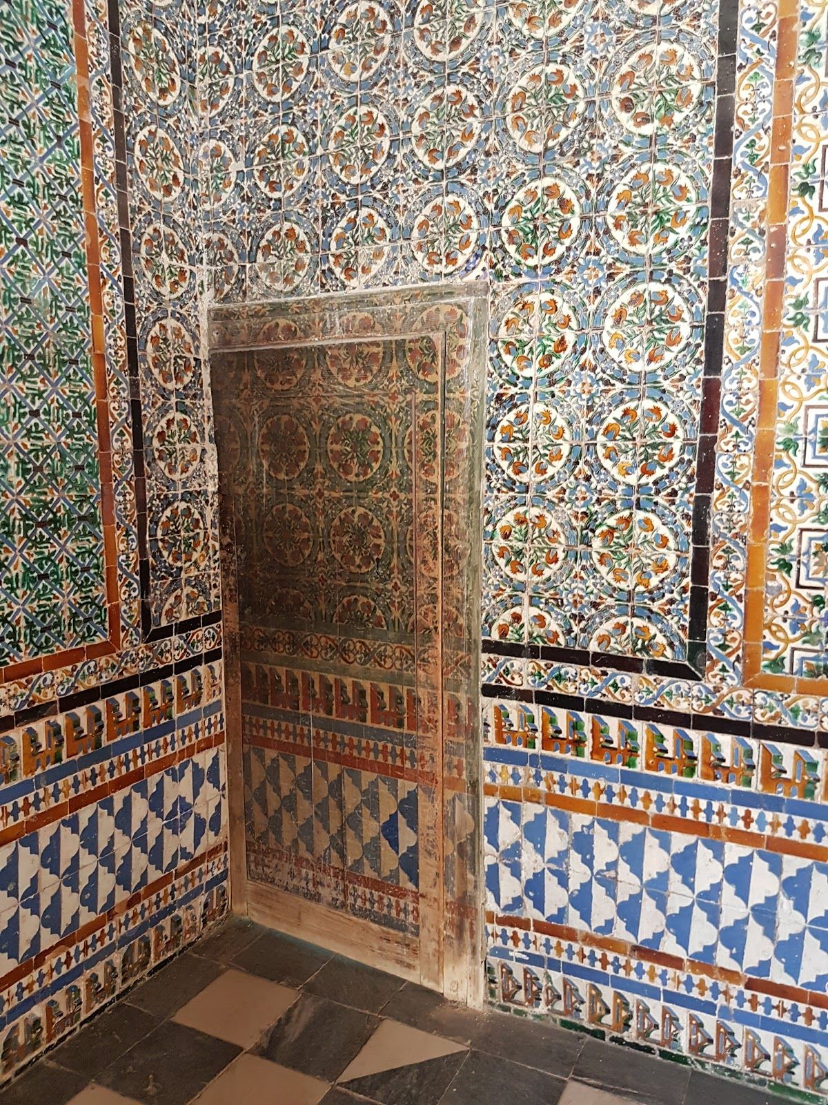 In a richly-tiled room corner, a wooden door is painted to look like the surrounding tile work, although age has darkened it to be more obvious.