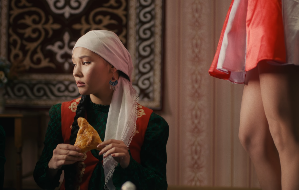 Image of a young woman dressed conservatively and wearing a headscarf, holding a samosa-like pastry, with the American in the stars-and-stripes dress standing mostly offscreen