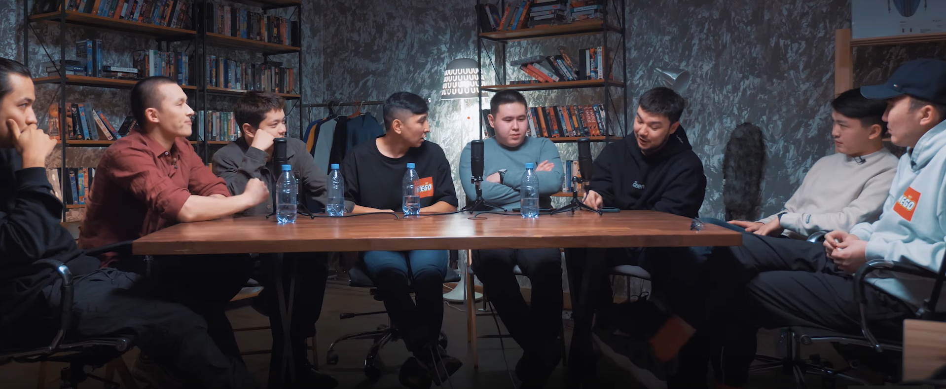 An image of seven men seated around a rectangular wooden table, with water bottles on the table and bookshelves and a lamp visible in the background