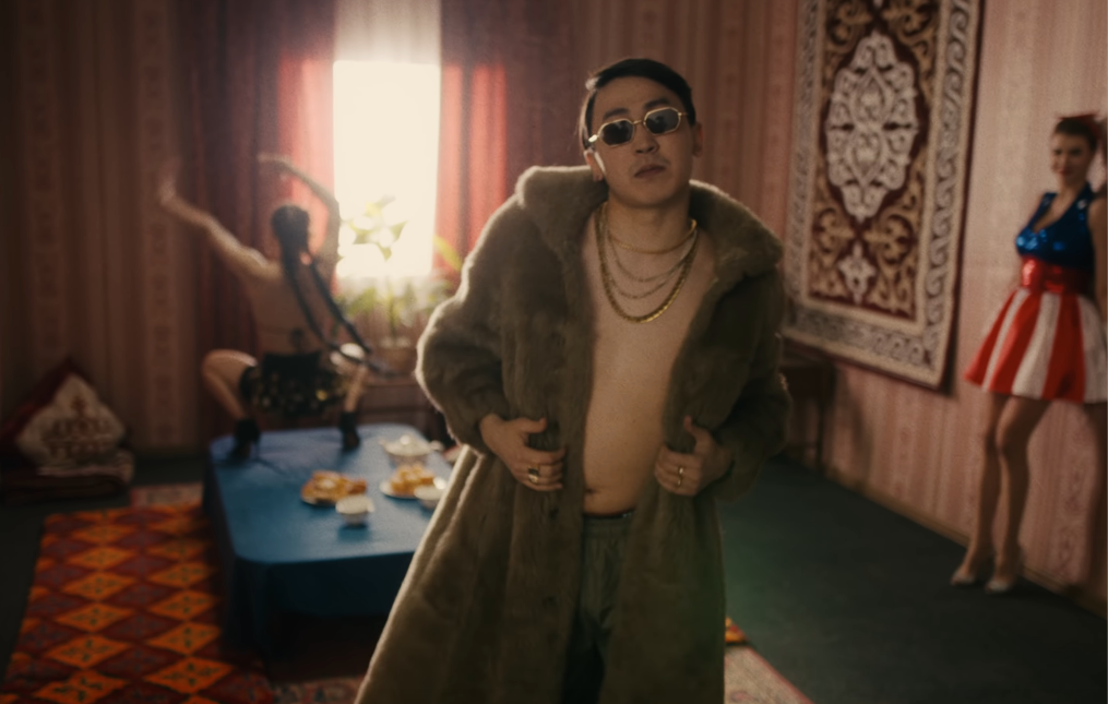 Image of Aldiyar wearing a knee-length fur coat, multiple gold necklaces, and sunglasses but no shirt as the dancer twerks in the background and the American in the stars-and-stripes dress looks on
