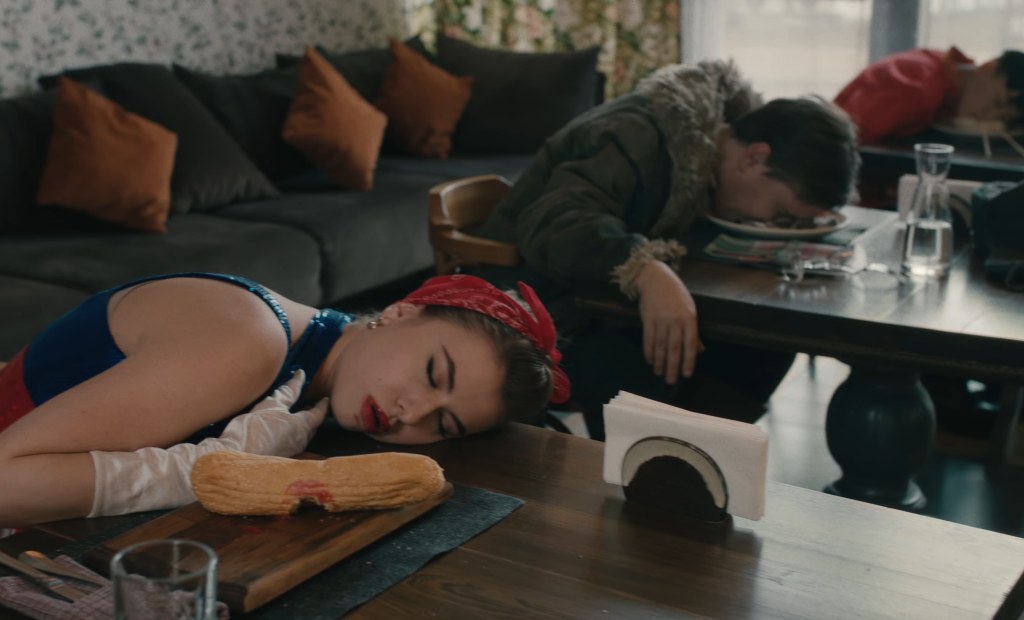 Image of three people in a restaurant, apparently dramatically dead, their heads in their food, one dressed in a stars-and-stripes dress, one in stereotypical Russian dress, one in stereotypical Chinese dress