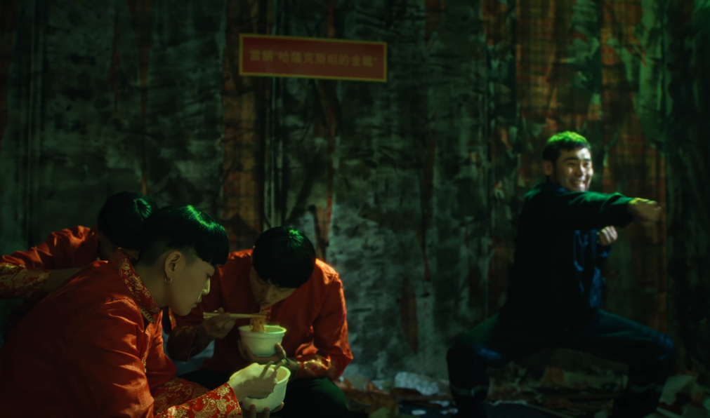 Image of Hiro in a blue outfit doing exercises and grimacing while the three people in bowl cuts eat noodles, with a sign in Chinese characters in the background