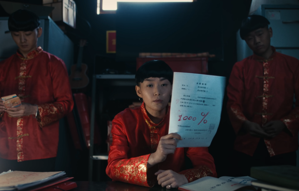 Image of three people in bowl cuts and red-and-gold jackets, one of them counting money and one of them holding up a piece of paper on which “1000%” is written large in red ink