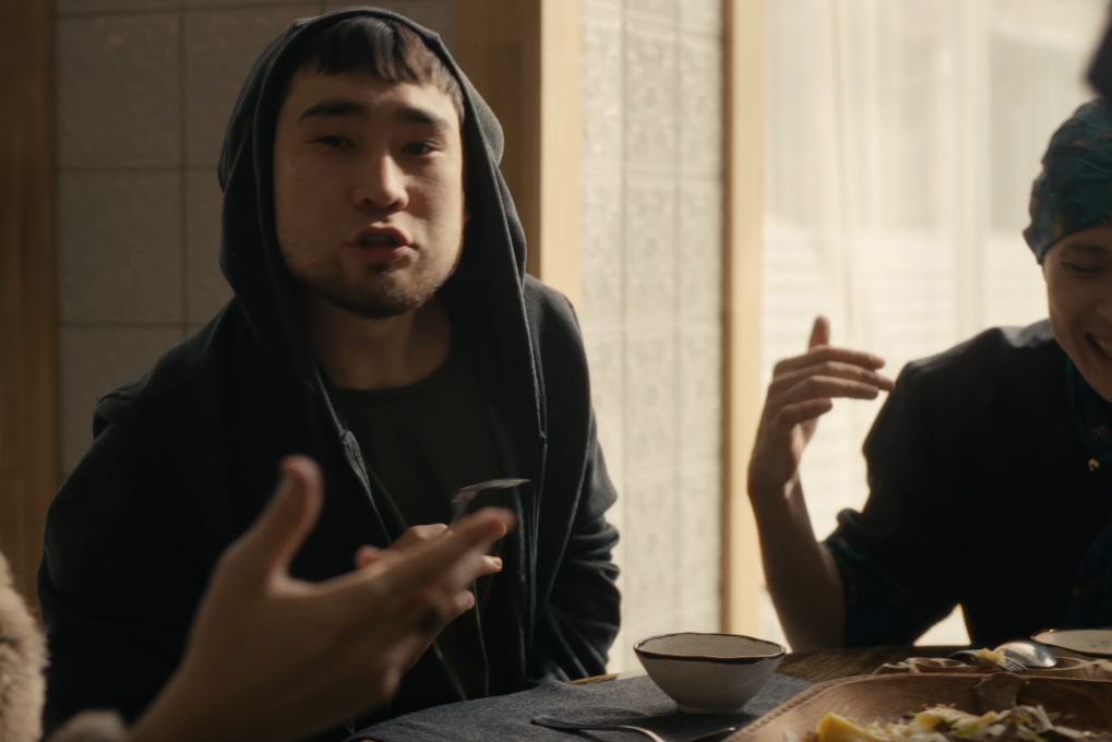 Image of Hiro sitting at a table, looking at the camera and rapping, De Lacure to his left