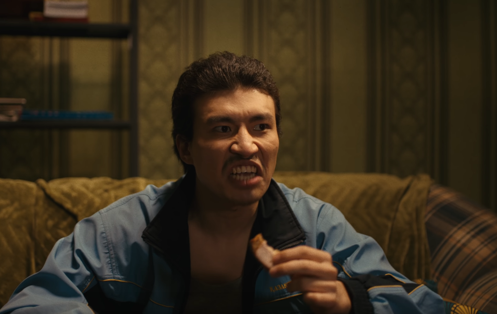 Image of Amazat dressed as the Kazakhstani equivalent of Archie Bunker, sitting on a couch while eating and looking angry