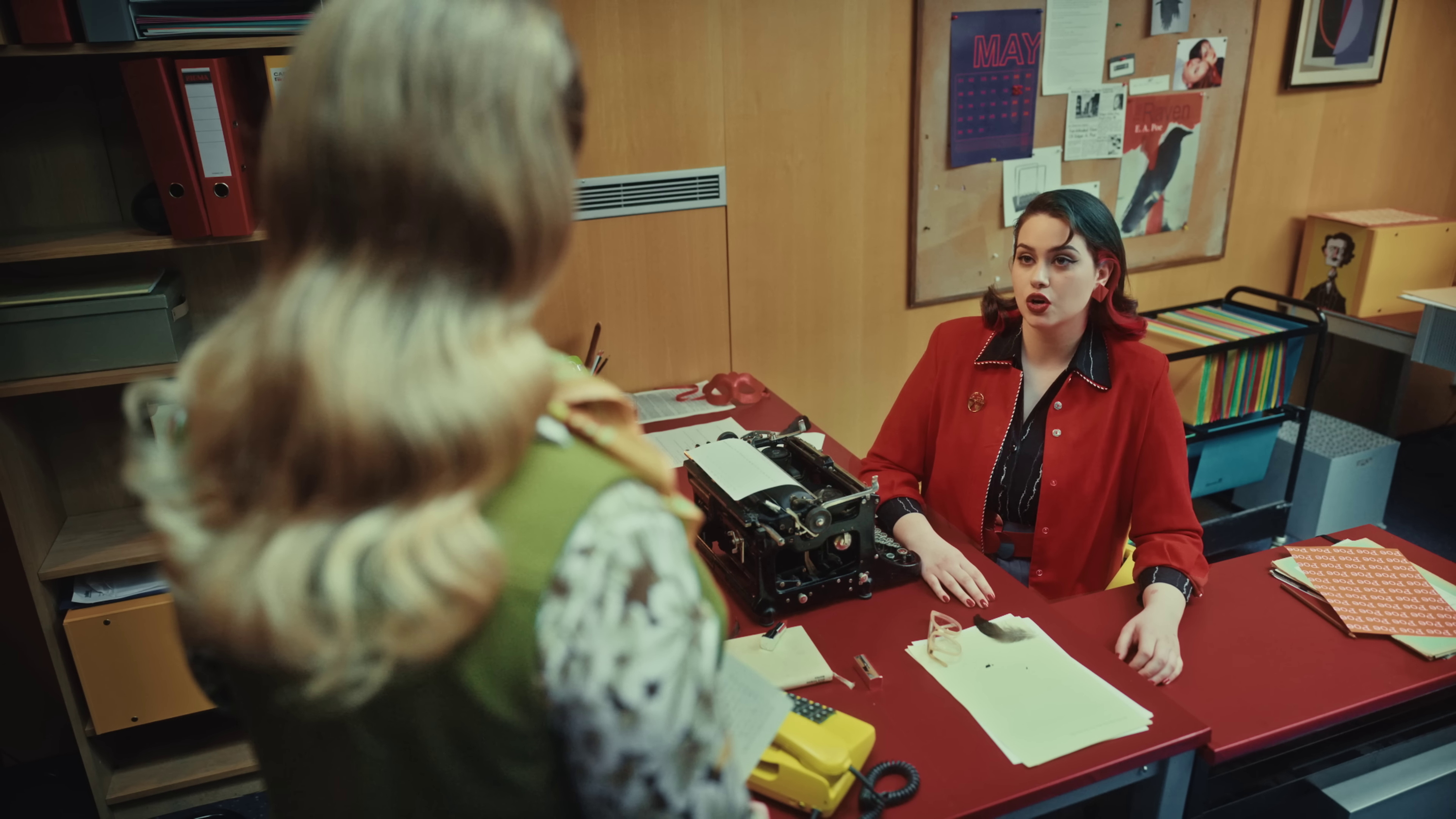 wide shot of Teya sitting at her desk with objects around her and a bulletin board behind her