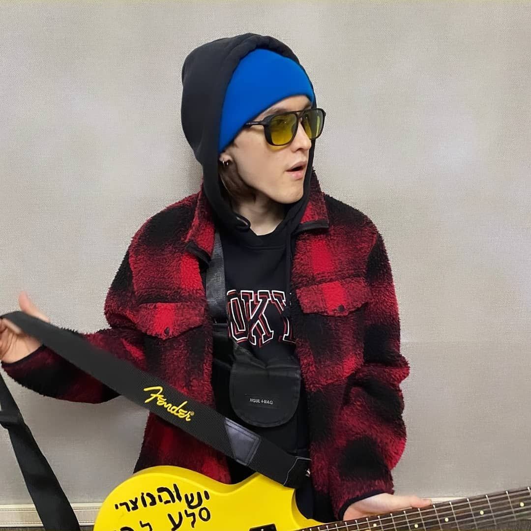 Image of Kyle Ruh looking off to the side while holding a yellow guitar with seeming Hebrew lettering on it, and wearing a hoodie and a jacket AND a hat AND sunglasses AND a small bag strapped to his chest