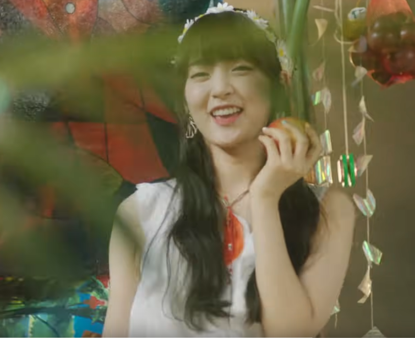 picture of Arin in a white dress and bangs, smiling at the camera while holding up an apple