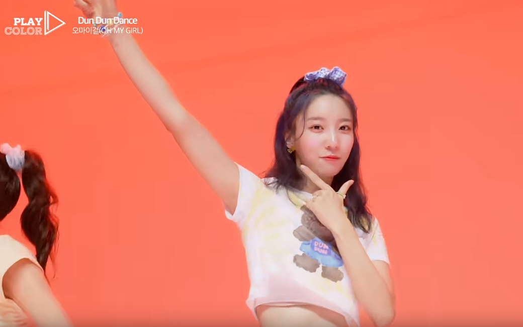 picture of Binnie wearing a small T-shirt against an orange background, with her right arm outstretched and her left hand under her chin