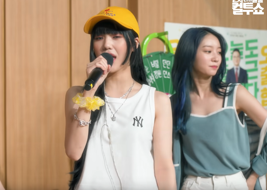 picture of Mimi in front, singing into a handheld microphone, while Binnie, behind her, looks off to the left