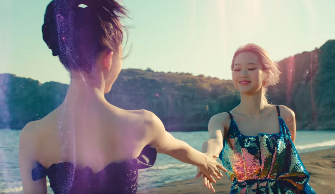 picture of Arin with her back to the camera, wearing a strapless dress, her hair pinned up, reaching out her right hand to Jiho, who is in the background, looking down and wearing a spangled dress with a seashell design