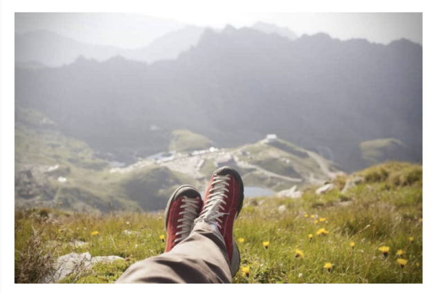 A person's feet on a grassy hill with mountains in the background Description automatically generated with medium confidence