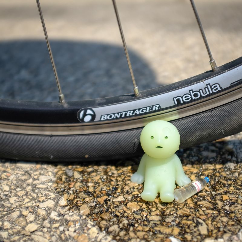 Small plastic Smiski figure sitting by a bicycle tire in a puddle of salty sweat