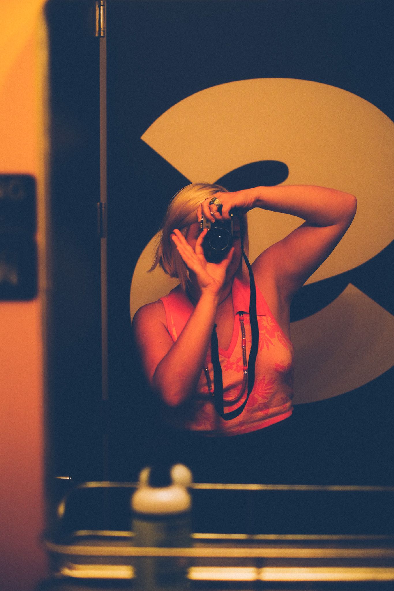 A self-portrait in a bar mirror, the photographer wearing a pink and orange tank top, face hidden by the camera.