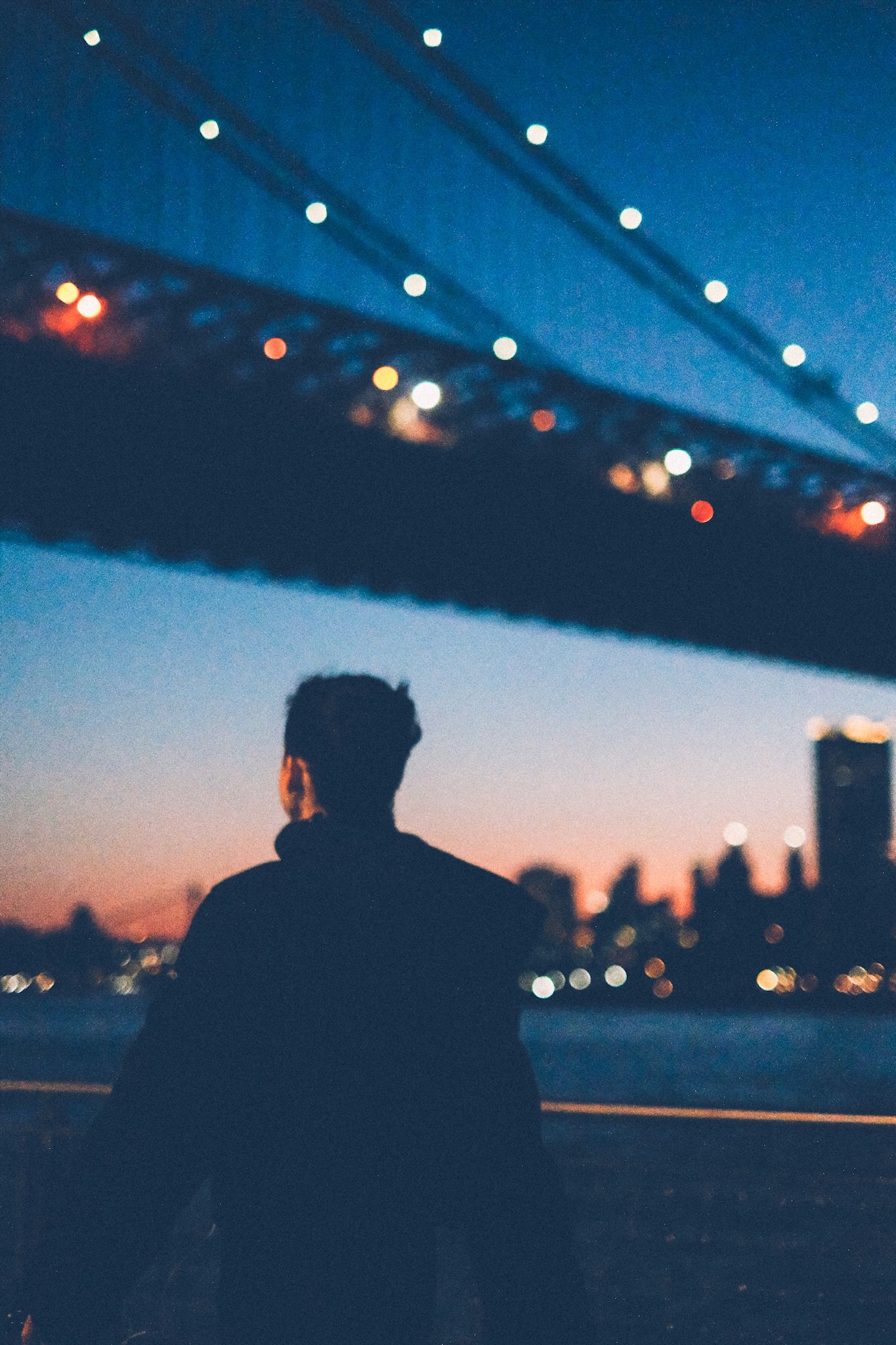 A person walks in front of the camera, the background showing the Williamsburg Bridge and the Manhattan skyline at sunset.