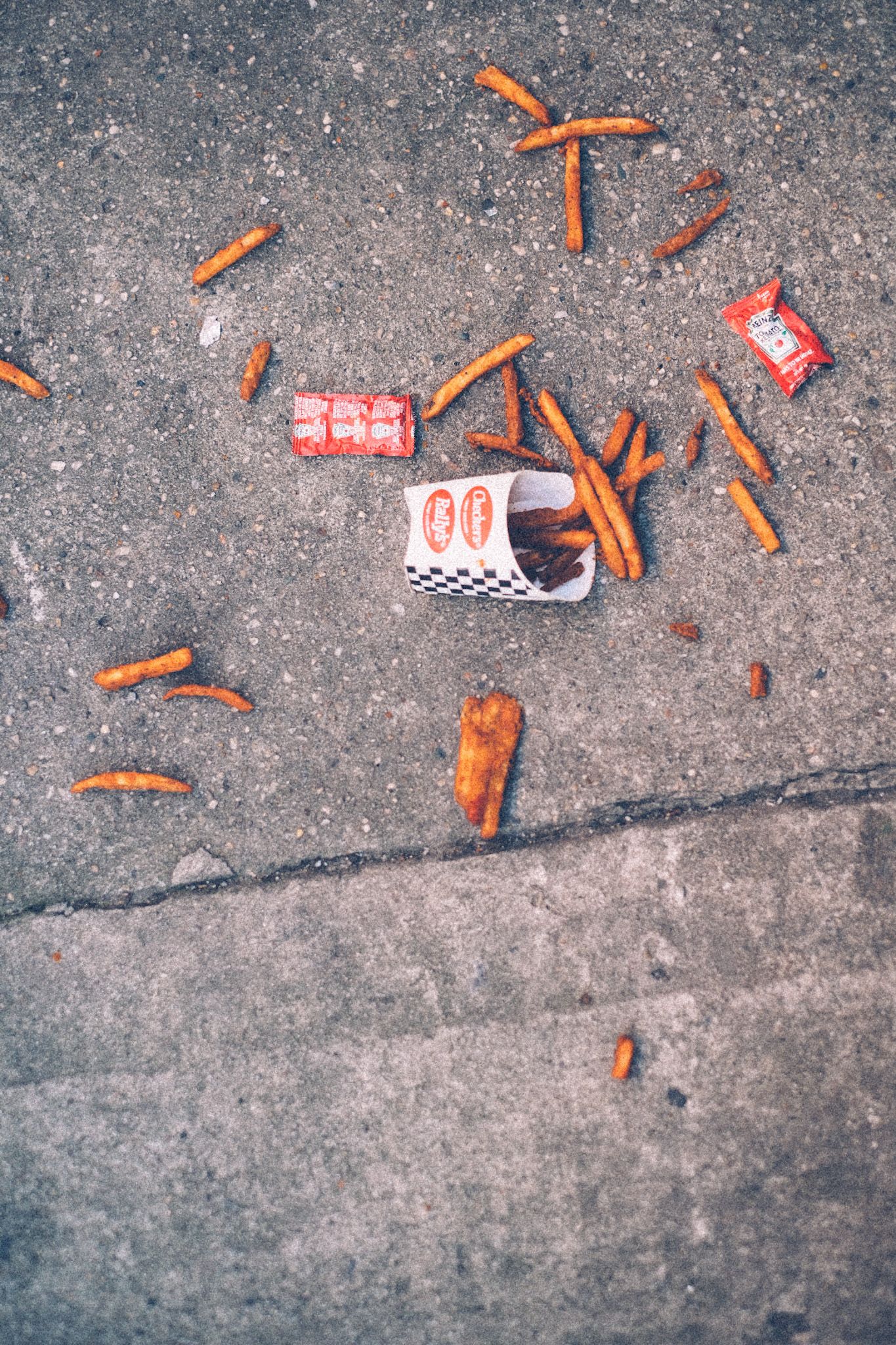 A sleeve of French fries has spilled on a concrete sidewalk, splaying out in all directions. Ketchup packets lie nearby. The black and white and red fry sleeve says Checkers.