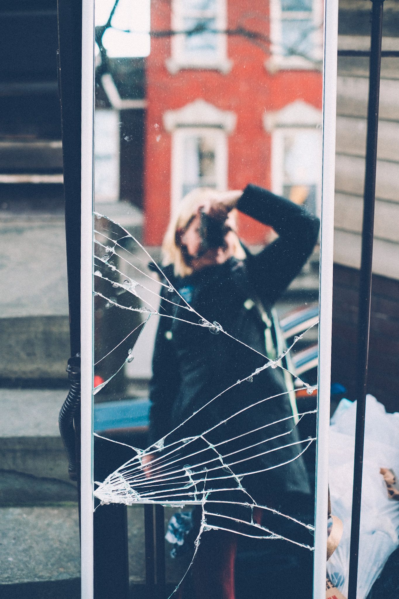 A self-portrait taken in a cracked mirror on a sidewalk, a red brick building in the reflection’s background.
