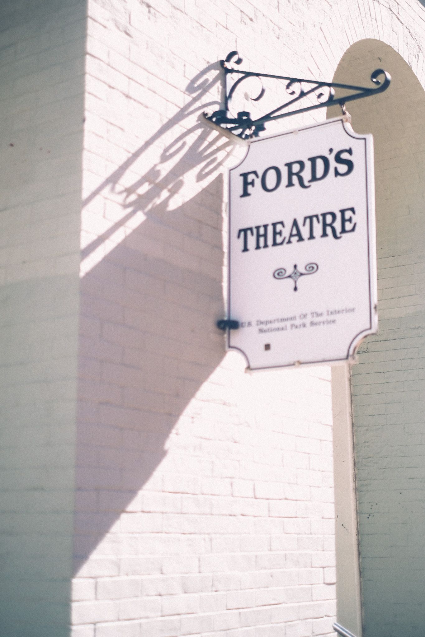 A white sign with black writing says “Ford’s Theatre,” attached to a white brick wall. Bright sunlight casts a shadow against the brick.