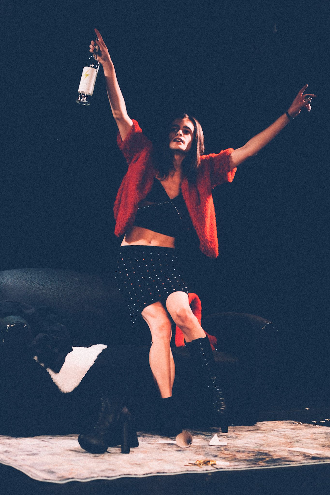 A stage actor is dressed in a black mini skirt and midriff with a furry red jacket and high-heeled boots, sitting on a couch while raising a bottle of vodka in the air.