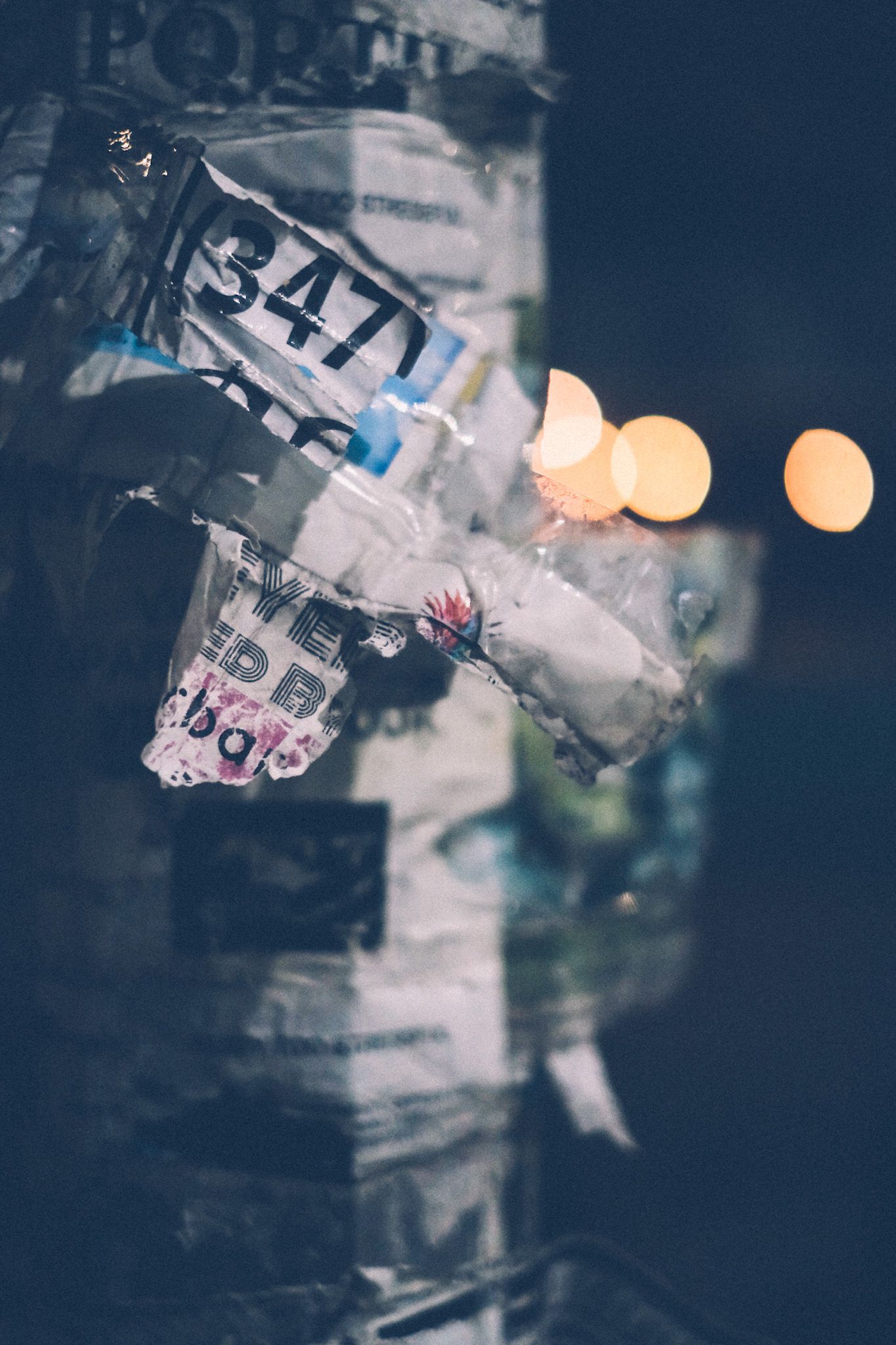 A bunch of different flyers have been papered on a light post. The main flyer, covered in tape, is ripped, but you can see the number 347 printed at the top, black on white. Lights bokeh in the background.
