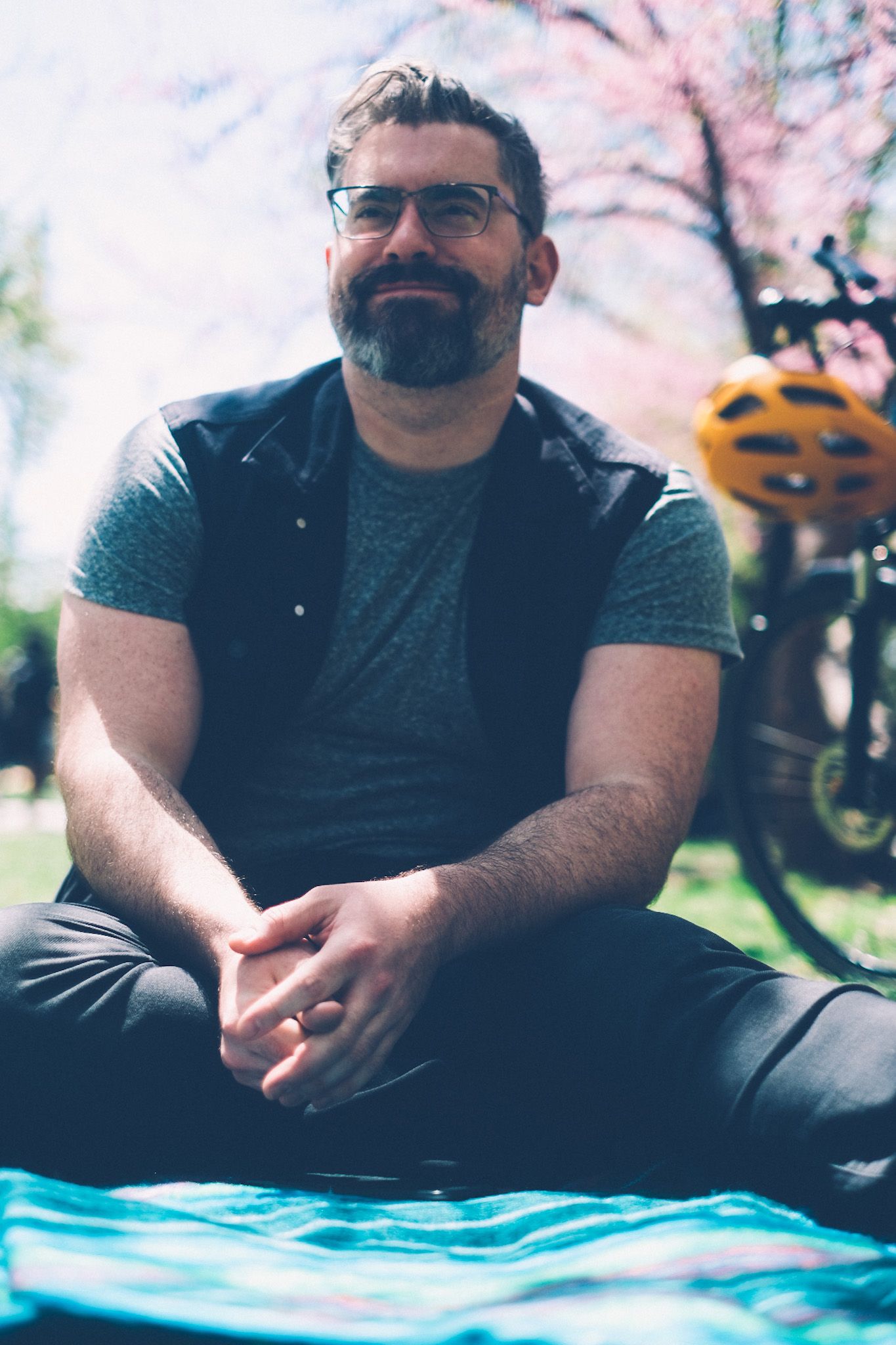 A man with dark hair, glasses, and a beard sits on a blanket in a park, smiling into the distance, his bike and helmet in the background against a cherry blossom tree.