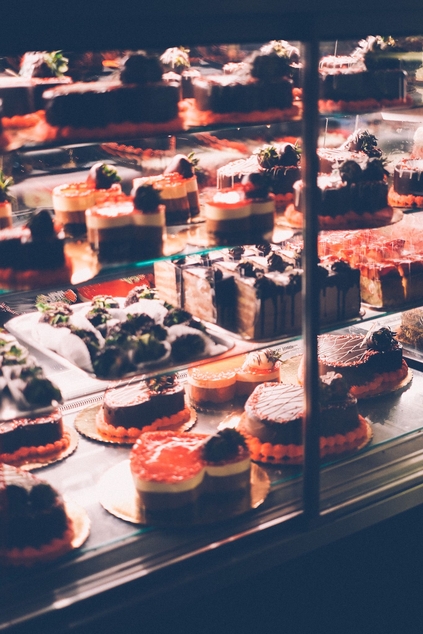Dozens of small and large heart-shaped cakes and other sweet treats of various flavors sit in a display case, decorated with chocolate and strawberries.