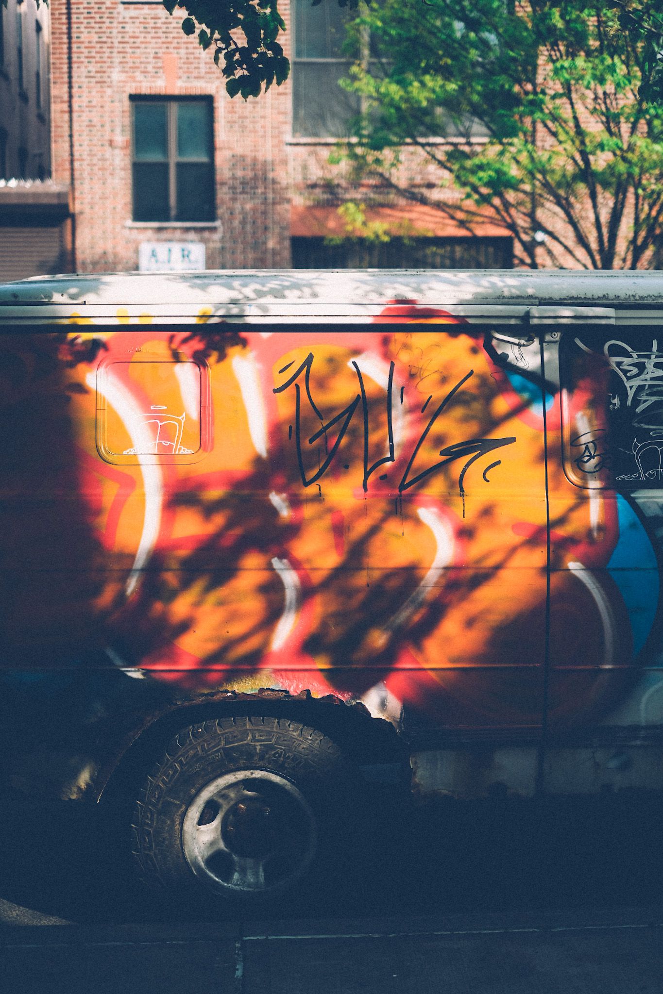 In the afternoon light, tree branch shadows fall over a van painted orange with pink and white graffiti.