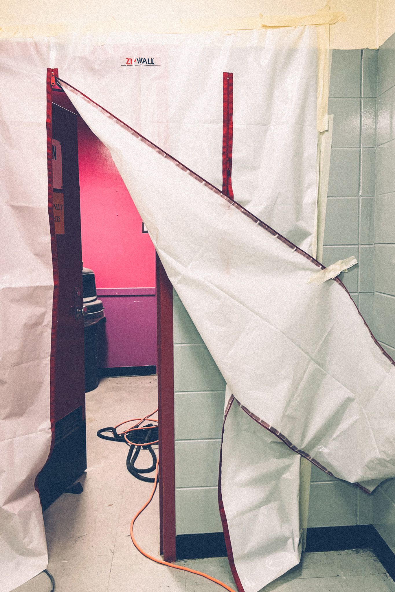 Clear construction tarp is taped across a doorway in a school, revealing a pink and purple wall in the back room.