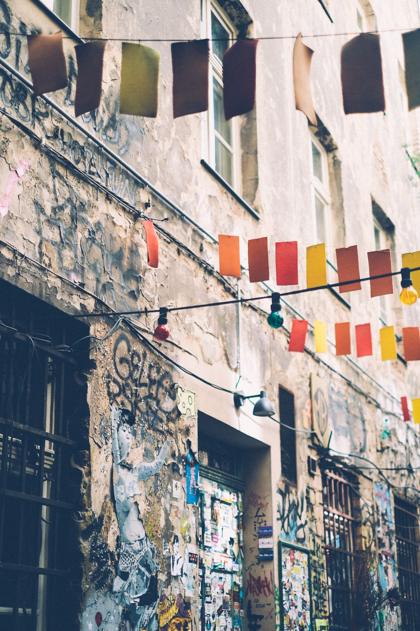 Little square pennant flags in warm colors hang in rows above a courtyard, the wall behind them filled with graffiti.