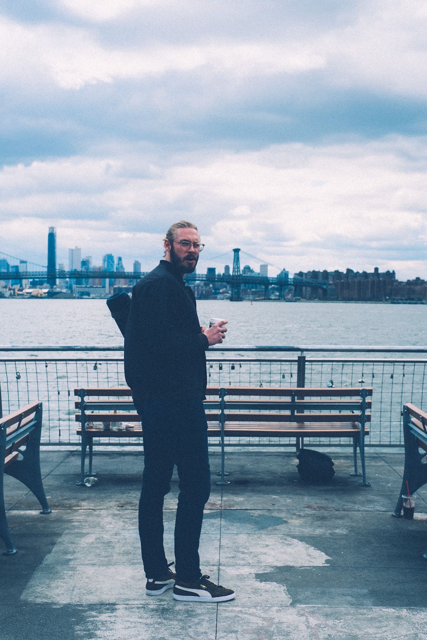 A man stands on a pier overlooking a bridge and the skyline of downtown Manhattan. It is a cloudy day. He holds a to-go cup of coffee and a yoga mat, turning to face the camera.