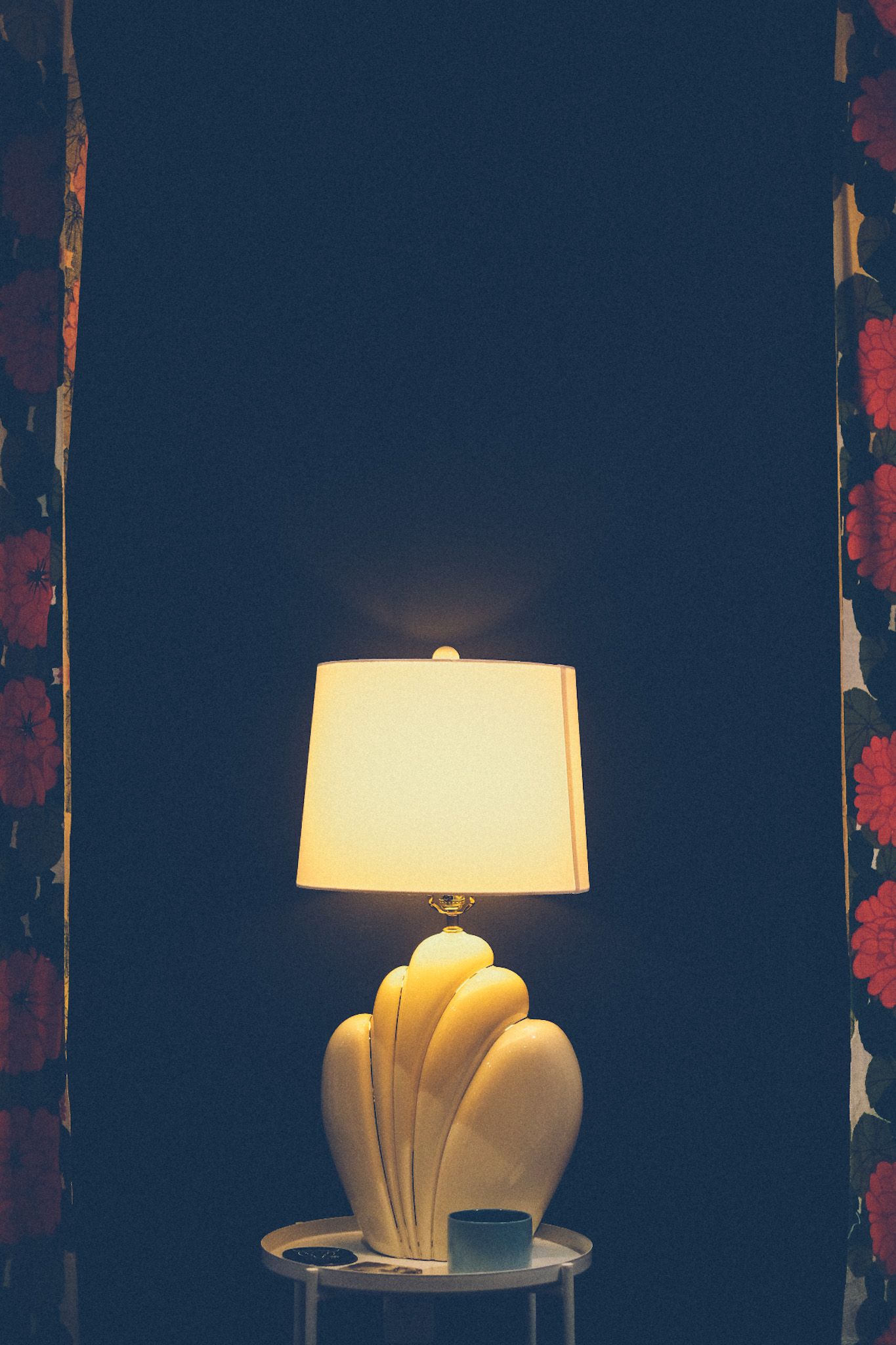 A cream-colored lamp from the 80s sits on a small table against a dark wall, framed by red floral curtains on either side.