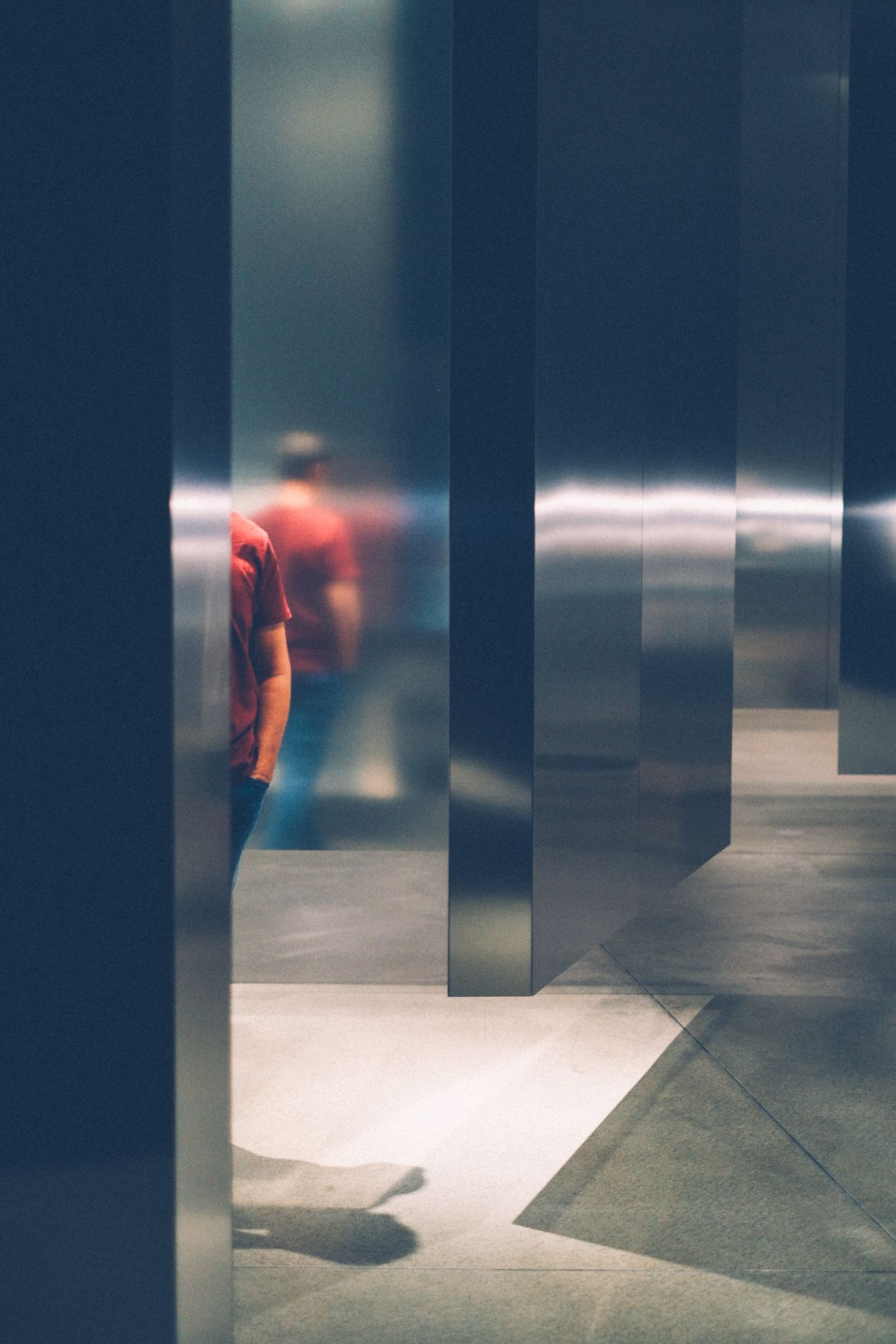 A slice of a torso of a man in a red shirt and jeans appears reflected among tall, aluminum, sharply angled columns.
