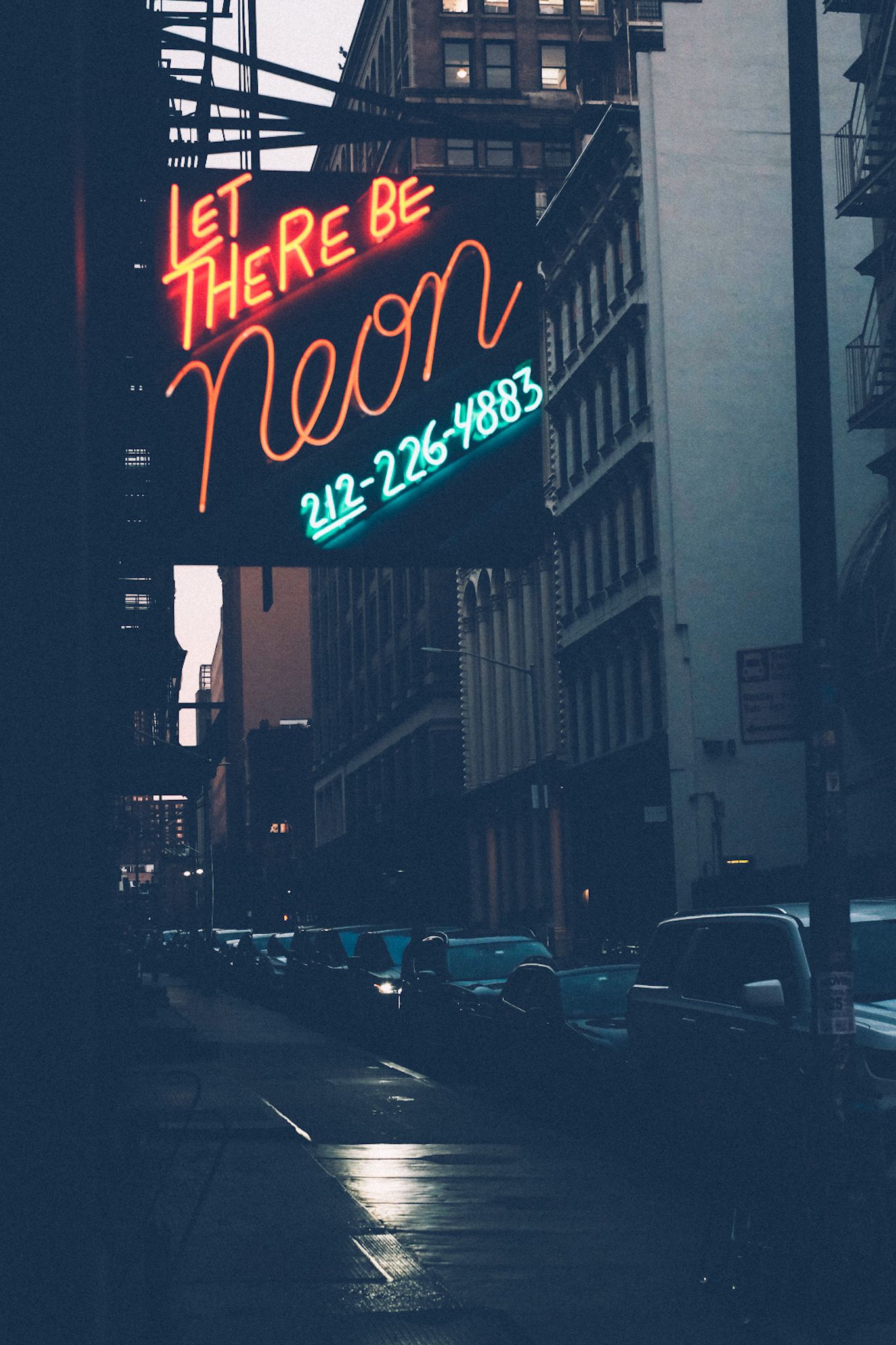 A multicolored neon sign saying “Let there be neon” hangs over a dreary Manhattan street.