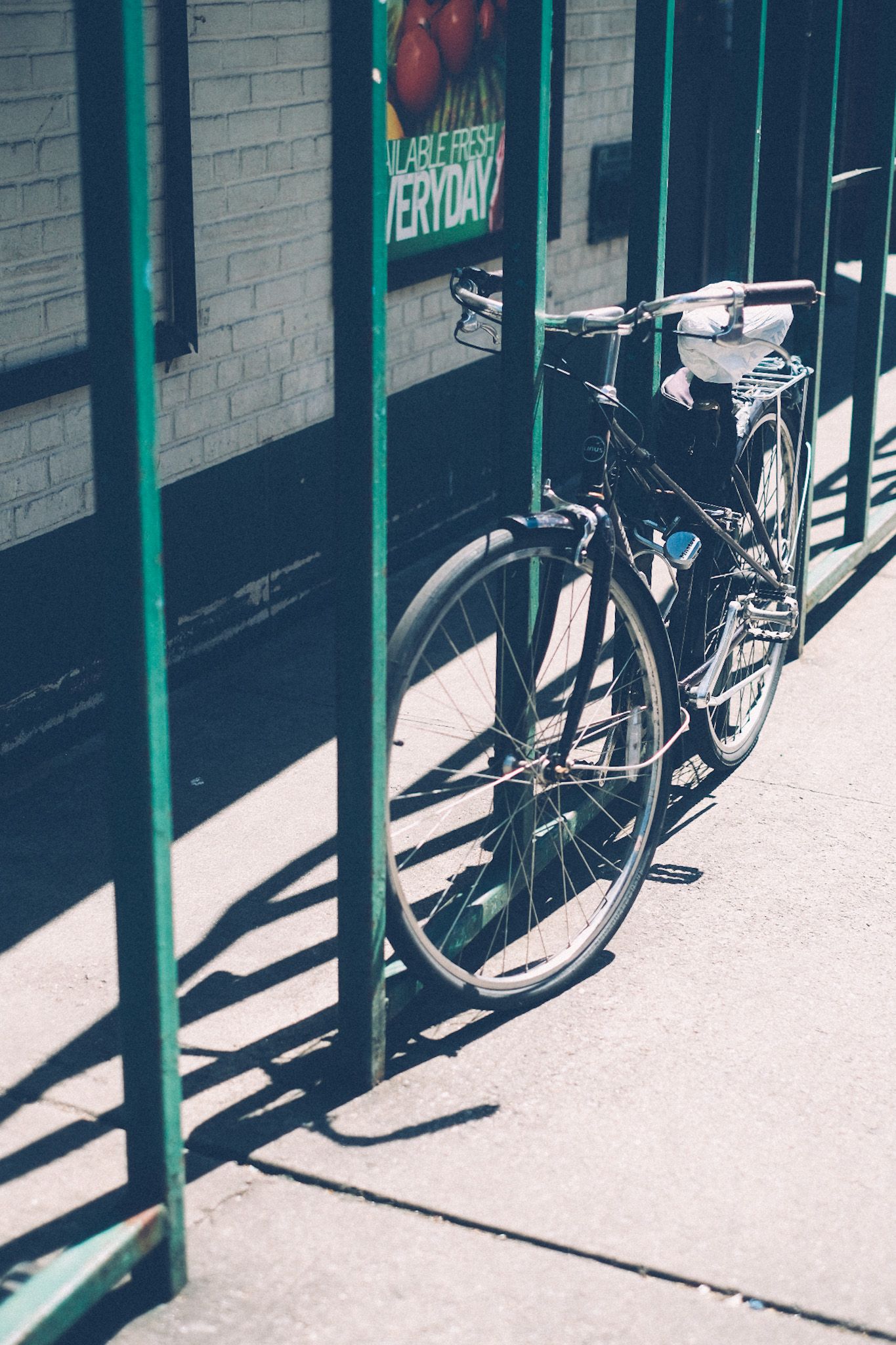 A bike is locked up against a green painted fence outside of a grocery store on a sunny afternoon.