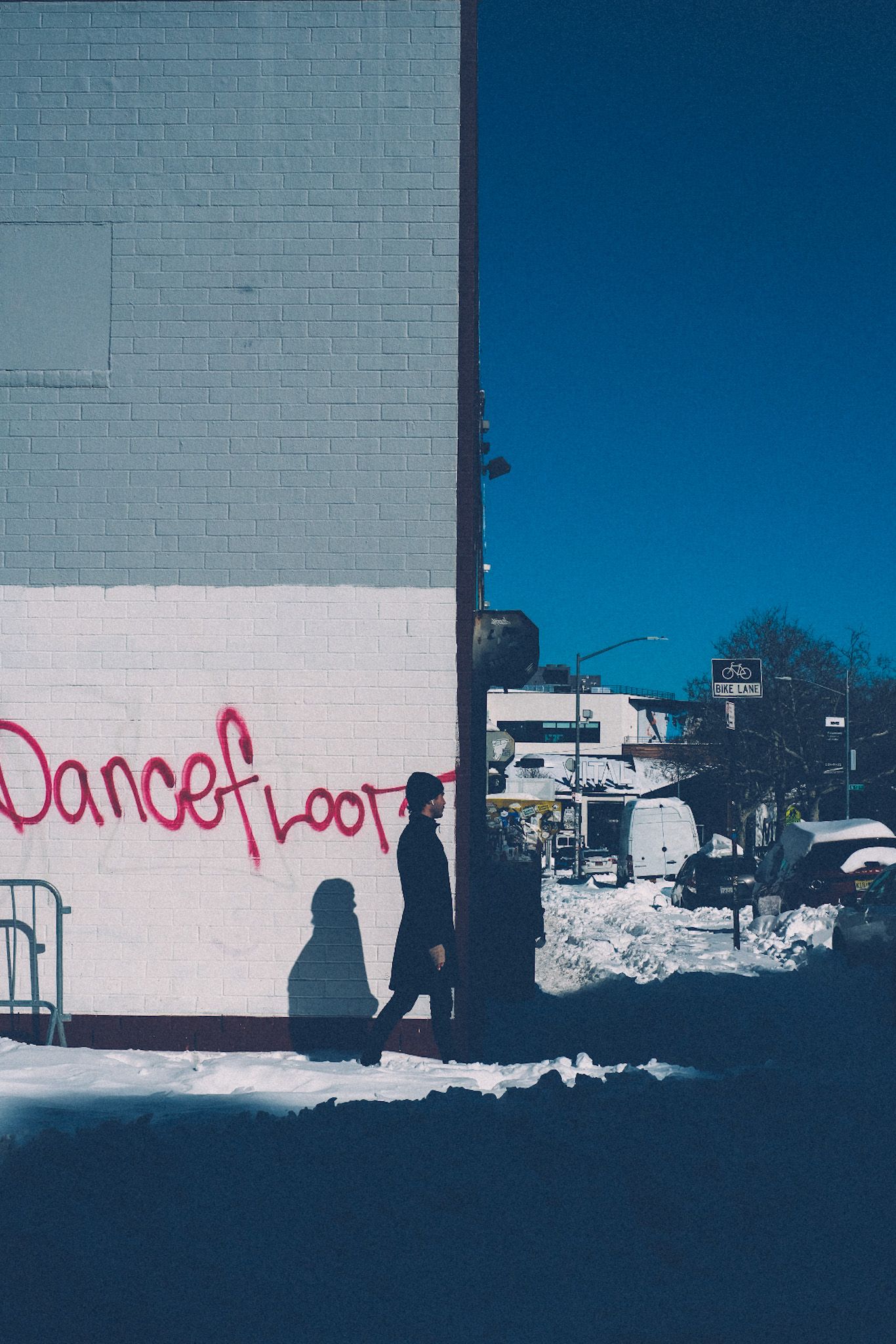 On a sunny day, piles of snow line the sidewalk where a person is passing in front of a building. The building, normally gray and white painted brick, displays the word “Dancefloor” in pink graffiti.