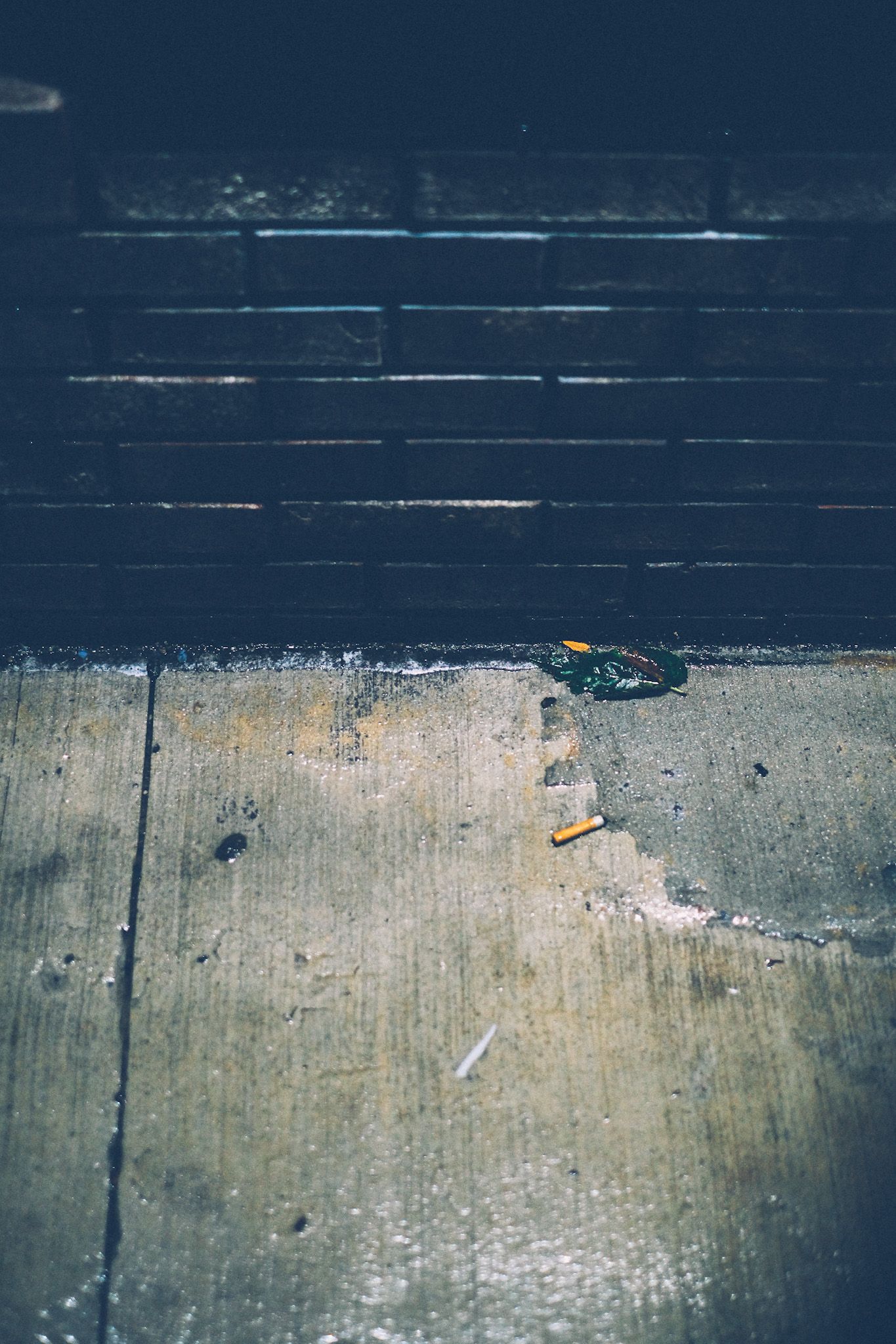 A cigarette sits on a wet sidewalk beside a brick wall at night, illuminated by street light.