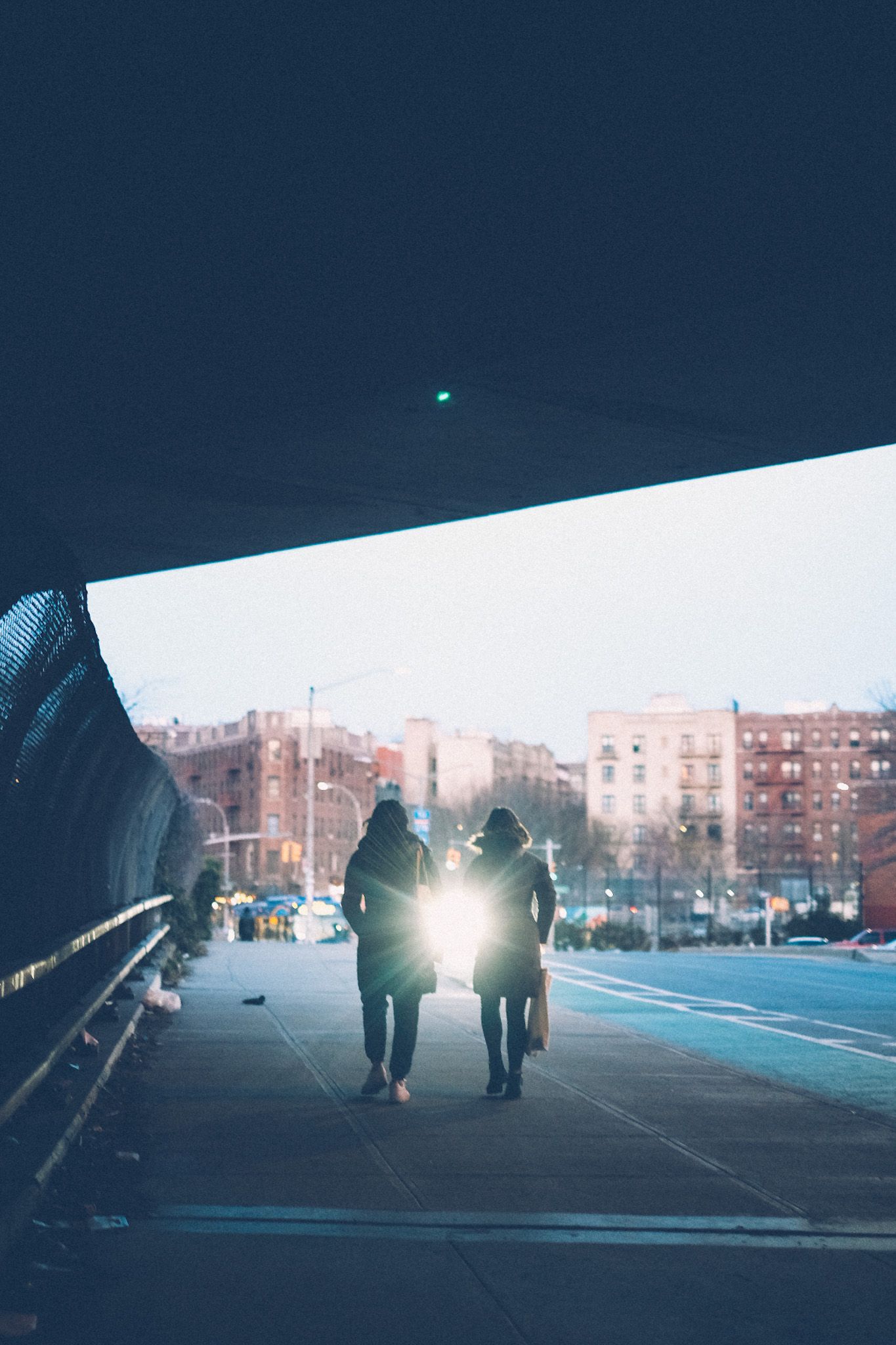 Two people walk under an overpass at dusk, a car headlight passing in the space between them.