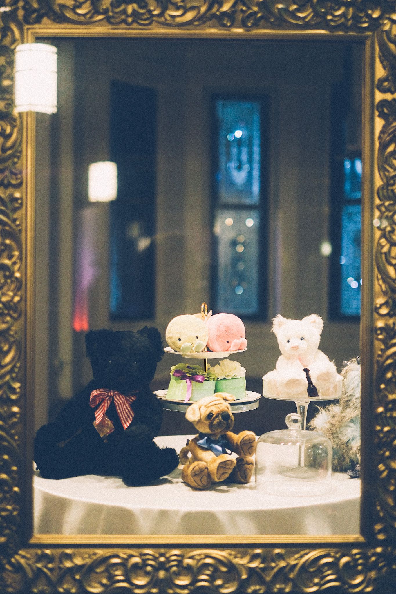 A few stuffed animals sit in the windowsill of an otherwise empty storefront, windows in the background showing night lights. A few of the stuffed things sit on a three tiered tea tray. An ornate gold frame is framing the picture.