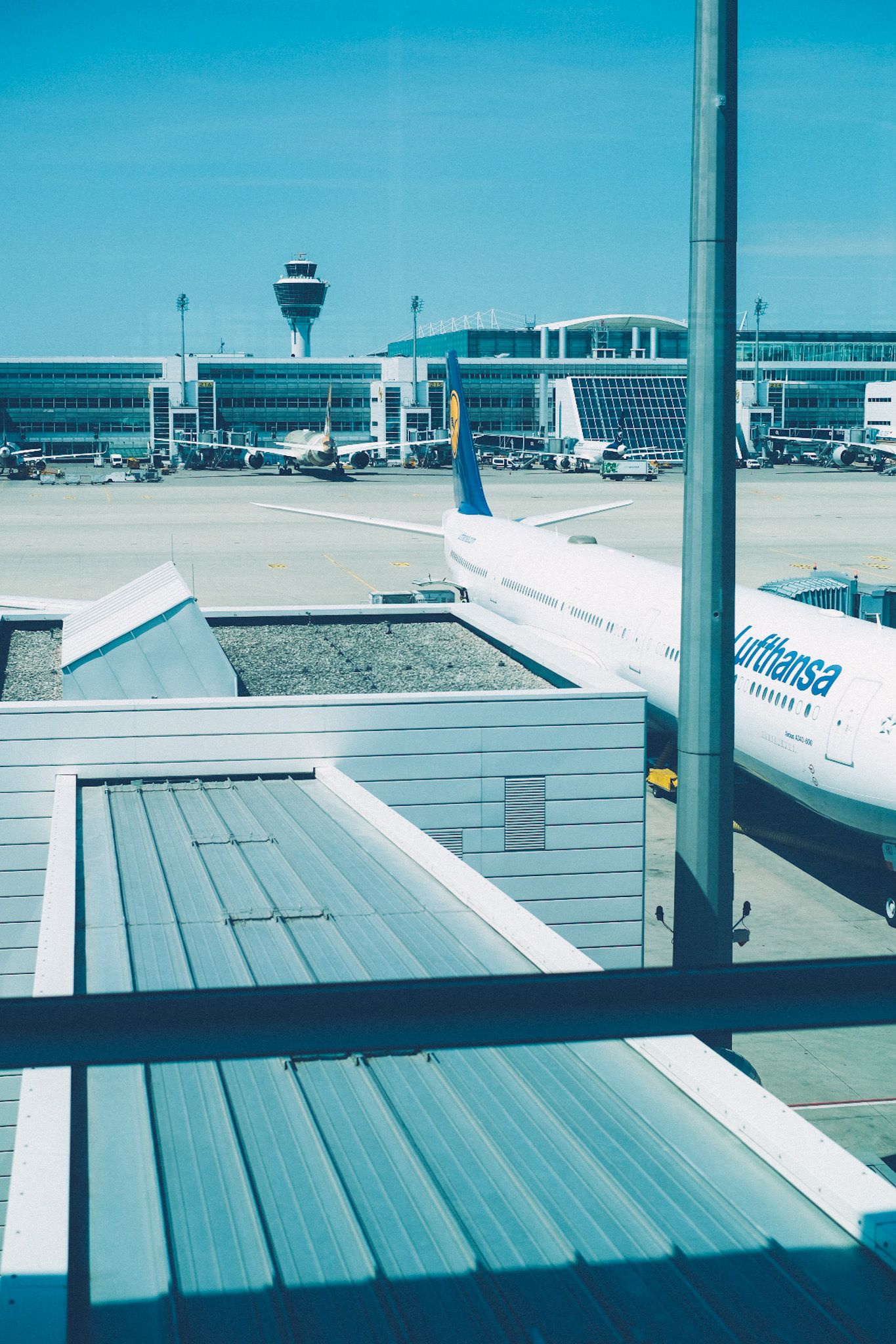 The view from a window of the Munich airport, staring out onto other terminals, grey pavement, a Lufthansa plane.