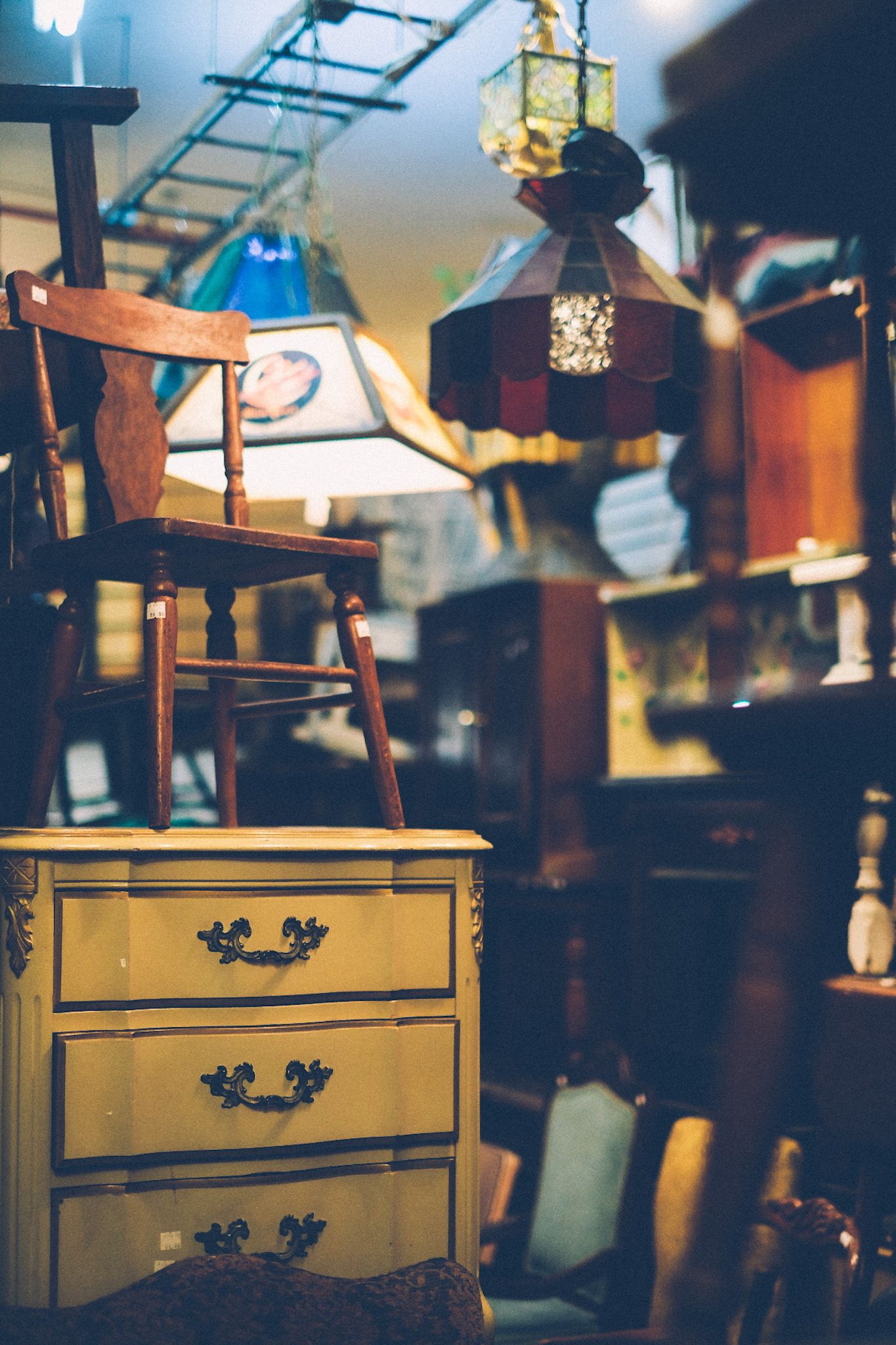 A wooden chair sits on top of a yellow dresser in an antique store. Lamps hang from the ceiling among all other clutter.