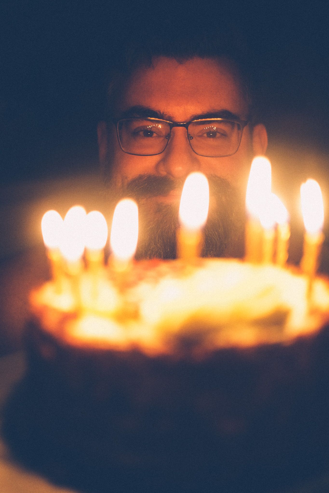 A bearded man with dark hair and glasses smiles from behind lit candles on a birthday cake, glowing, warmly lit.