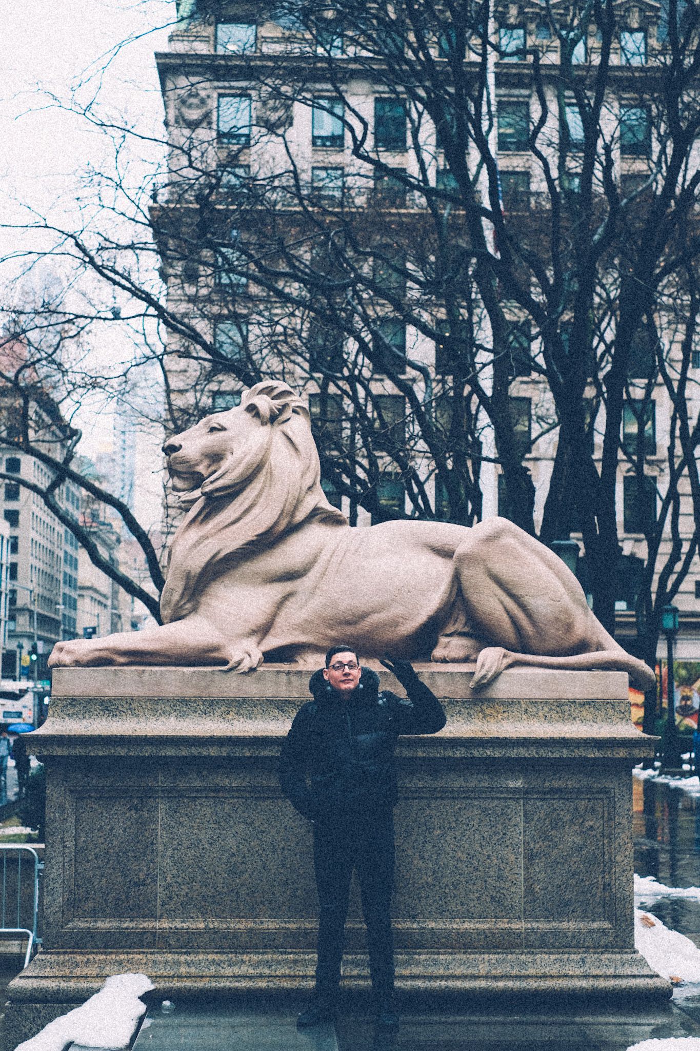 A man in all black stands in front of a gigantic lion statue, the ones outside of the New York Public Library�s main branch. Small mounds of snow and water puddles surround him.