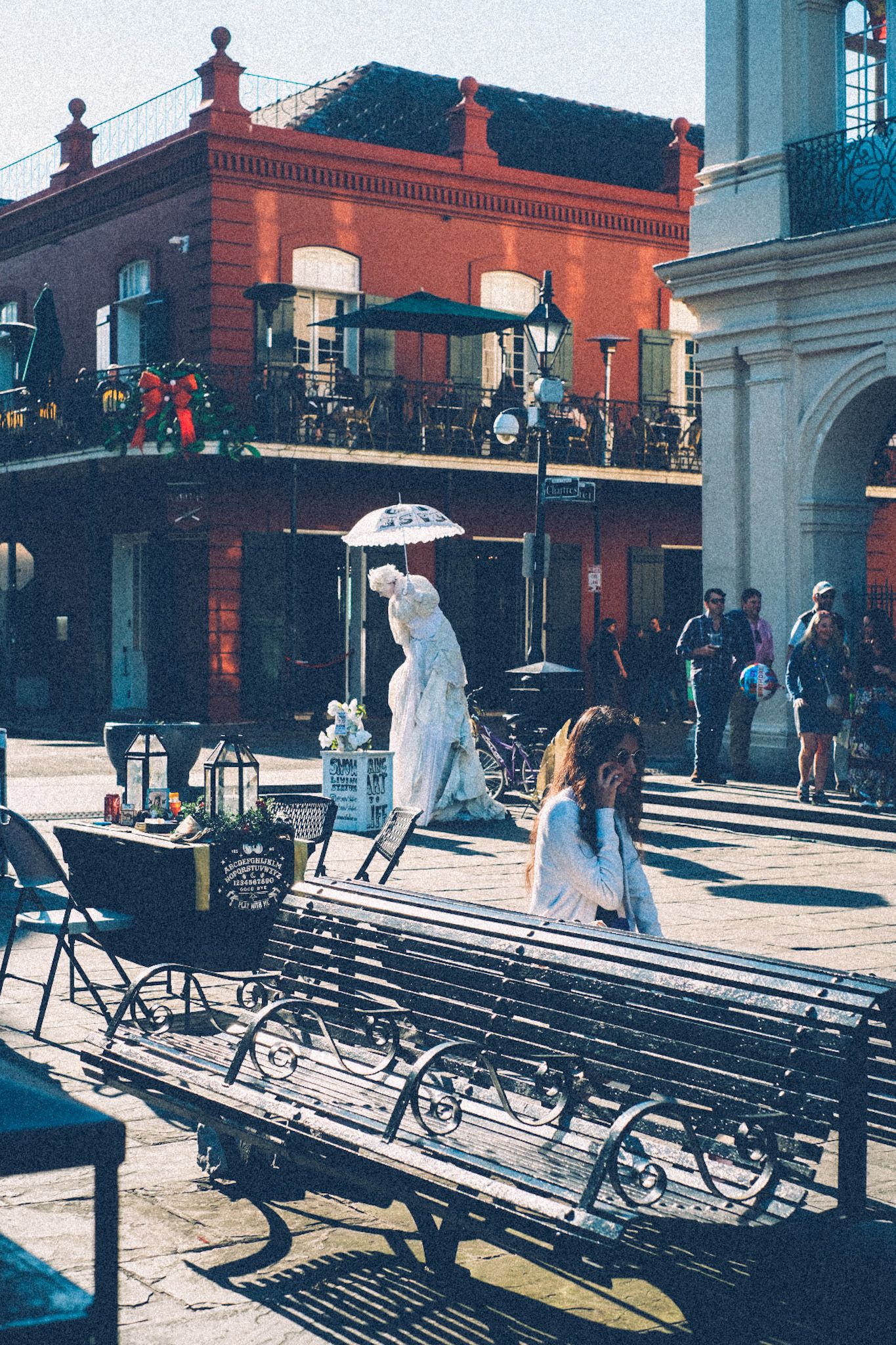 A woman posing as a tall Victorian statue covered in white paint stands in the background, among red brick buildings in the French quarter.