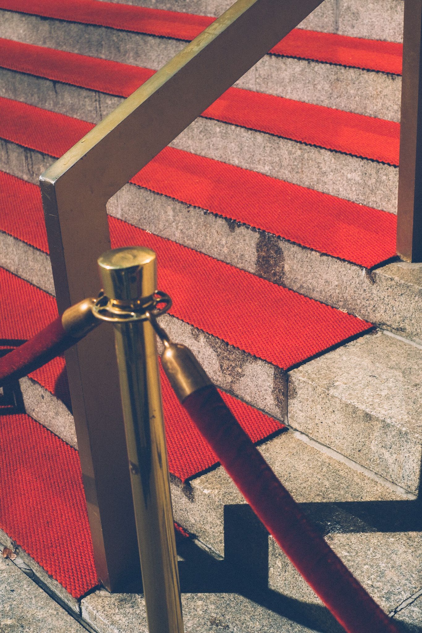 A velvet rope hung from a golden stand lines red carpeted stairs outside of a building.