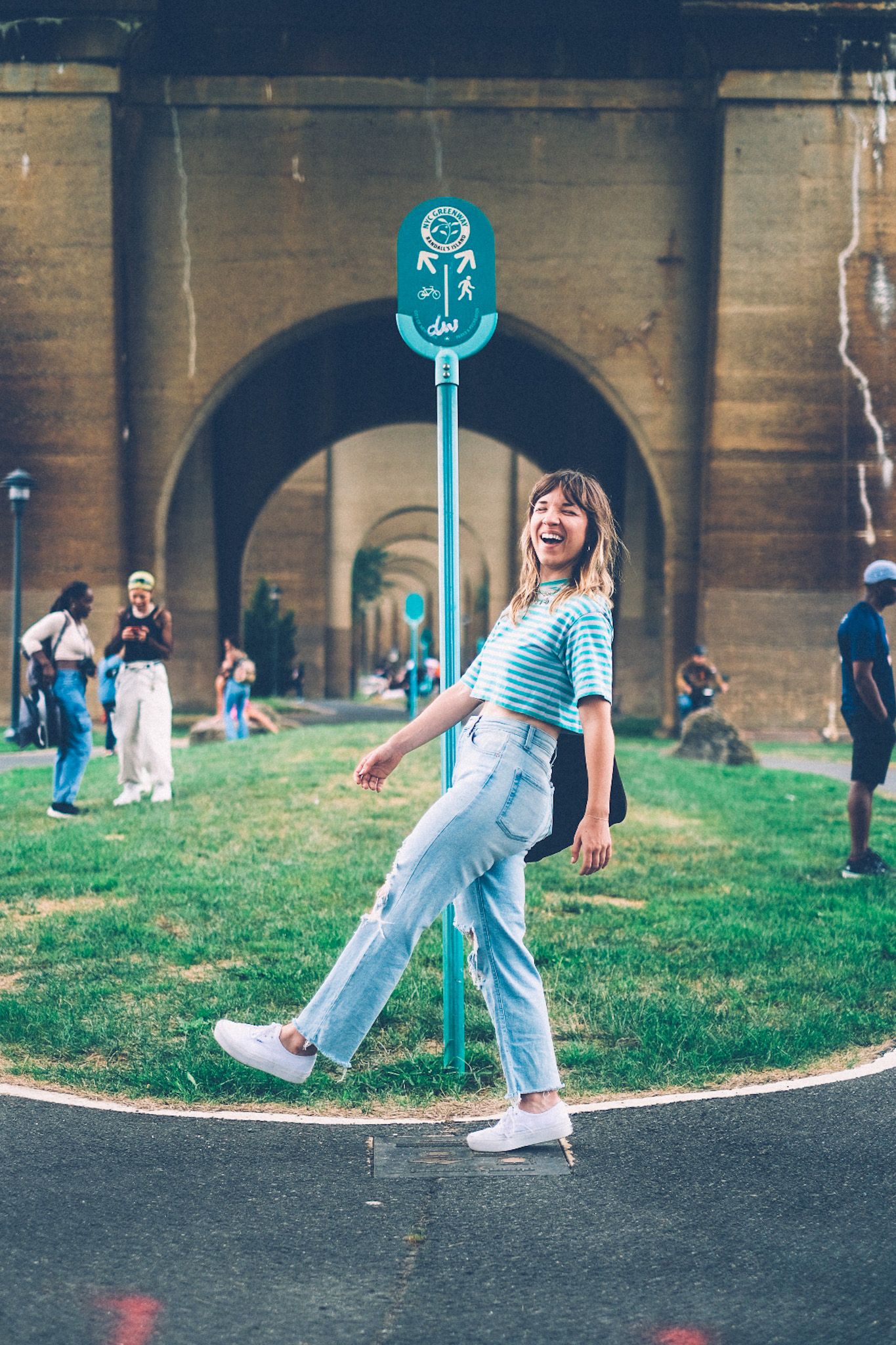 A woman kicks her leg out in front of her while standing in front of a turquoise pedestrian sign, laughing in front of arches of a bridge in the background.