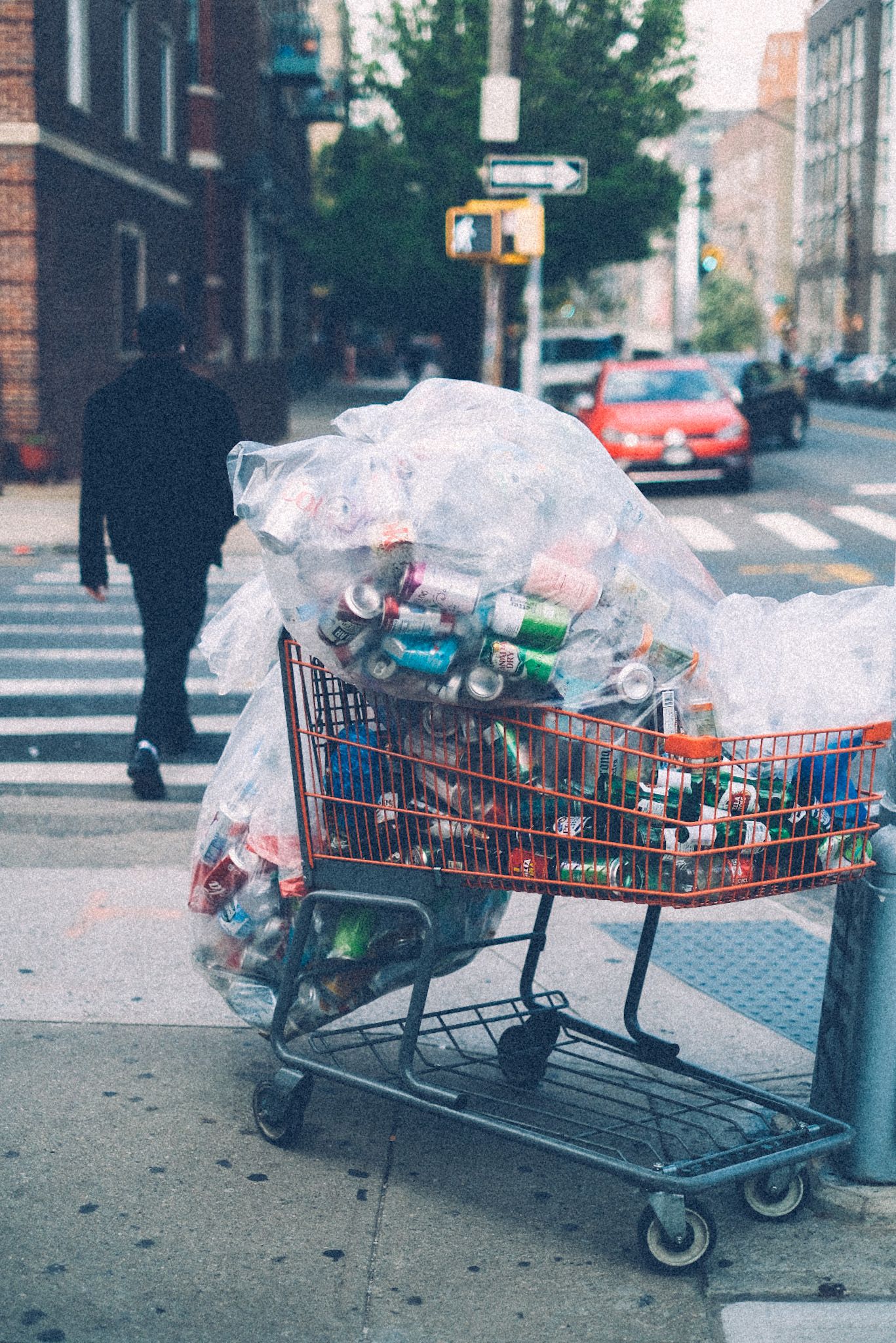A man in the background walks past a shopping cart full of recycled soda cans on the corner of a sidewalk.