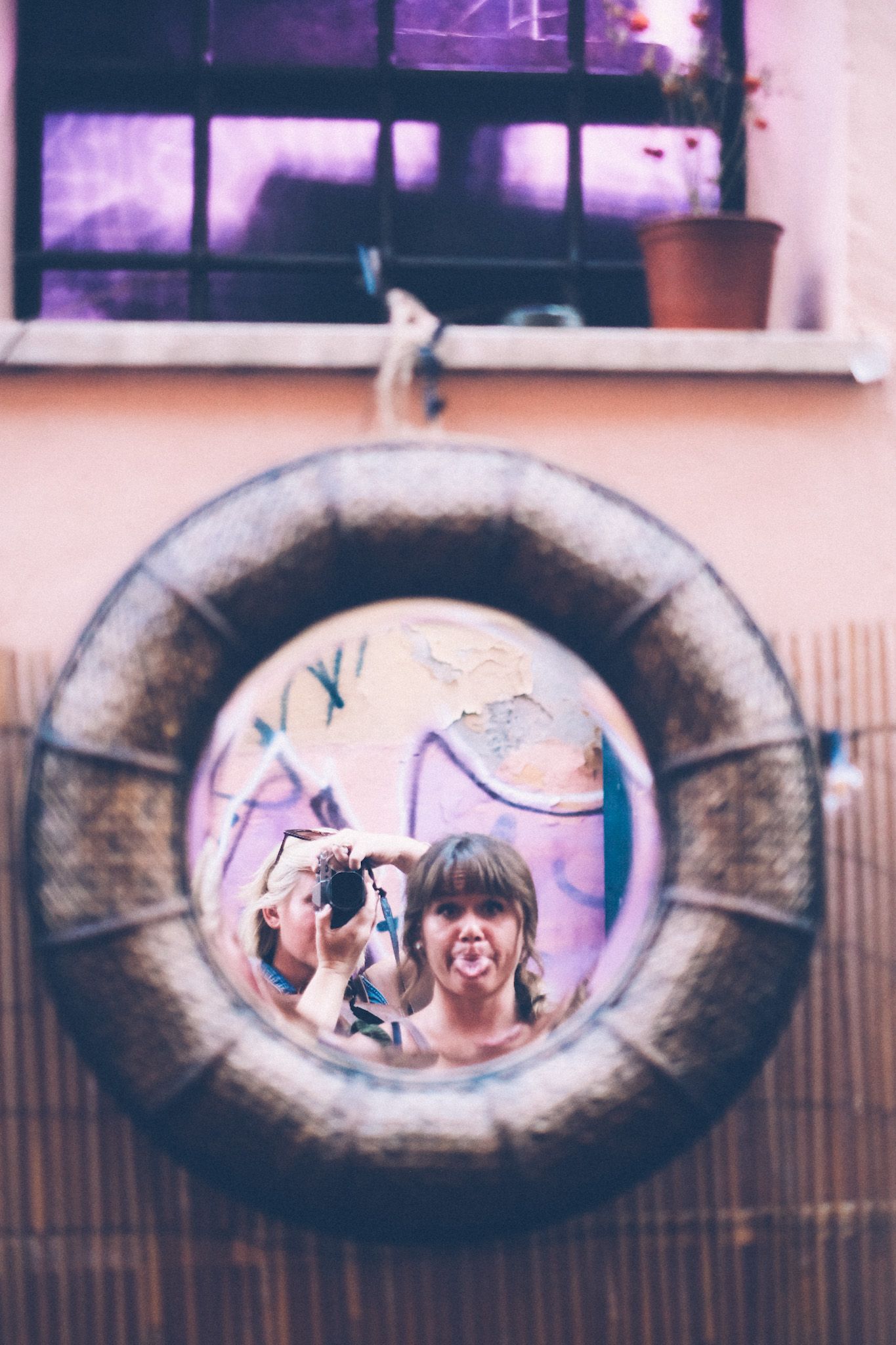 Two women look into a round mirror that looks like a porthole underneath a purple glass window, graffiti in the reflection. The woman in front is making a silly face.