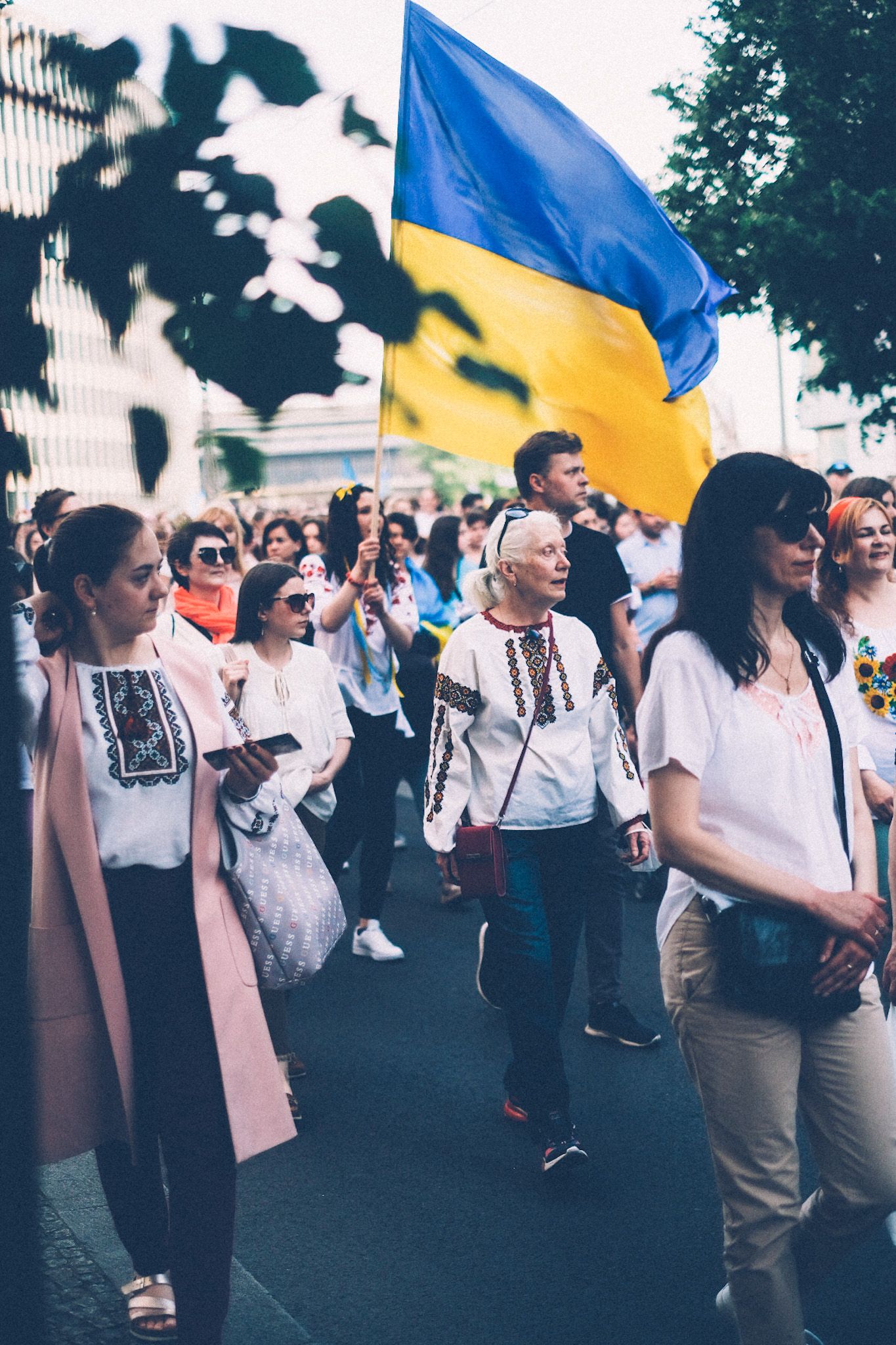 A crowd of protestors walk down the city street, a huge Ukrainian flag waving across the background.