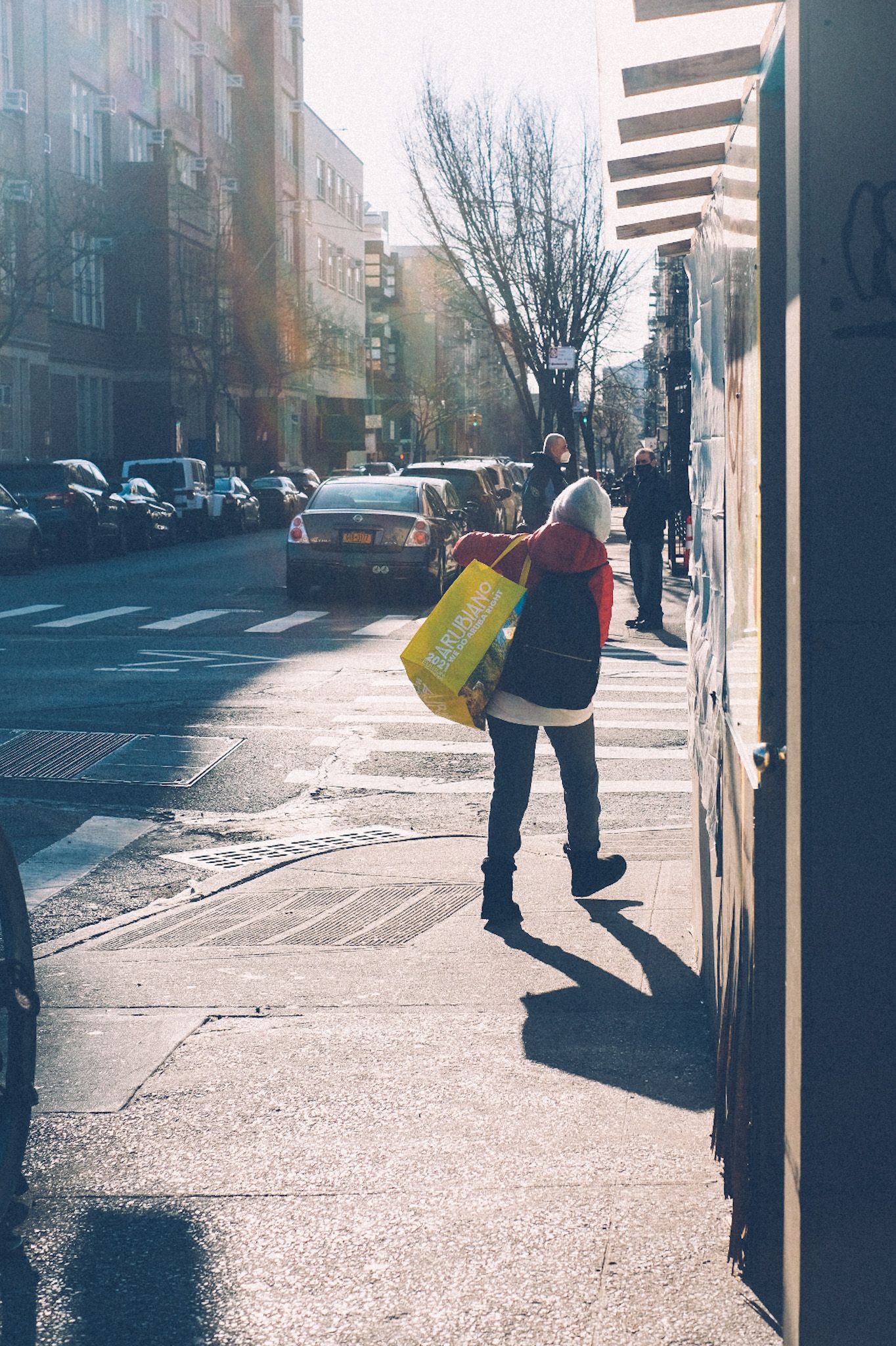 A woman carries a grocery tote down the street in the morning light, two men standing in the background looking.