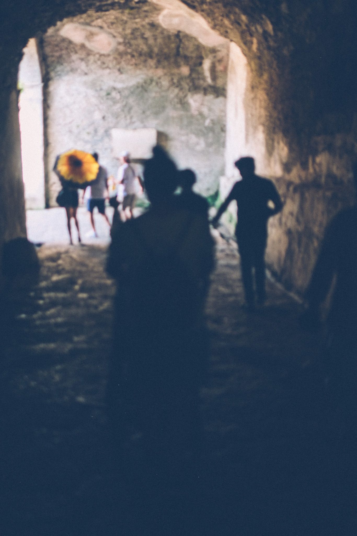 Shadowy, unfocused silhouettes walk through a tunnel into the light of the colosseum at Pompeii.